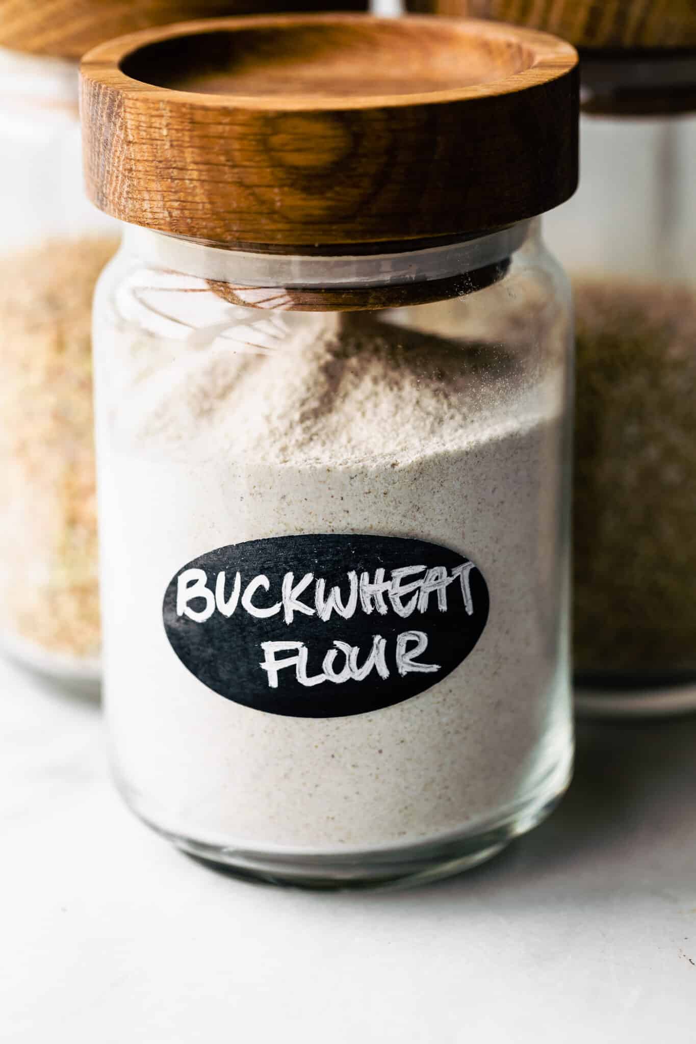 A clear glass jar of buckwheat flour with a wooden lid and black label.