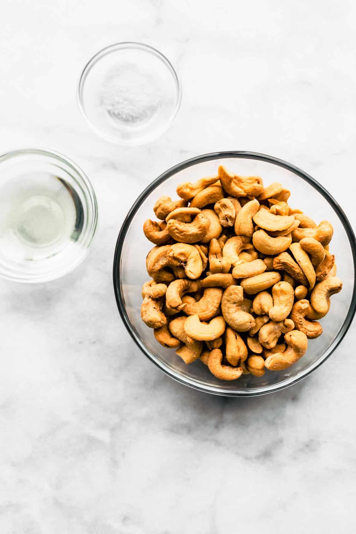 Separate bowls of raw cashews, coconut oil, and salt on a marble countertop.