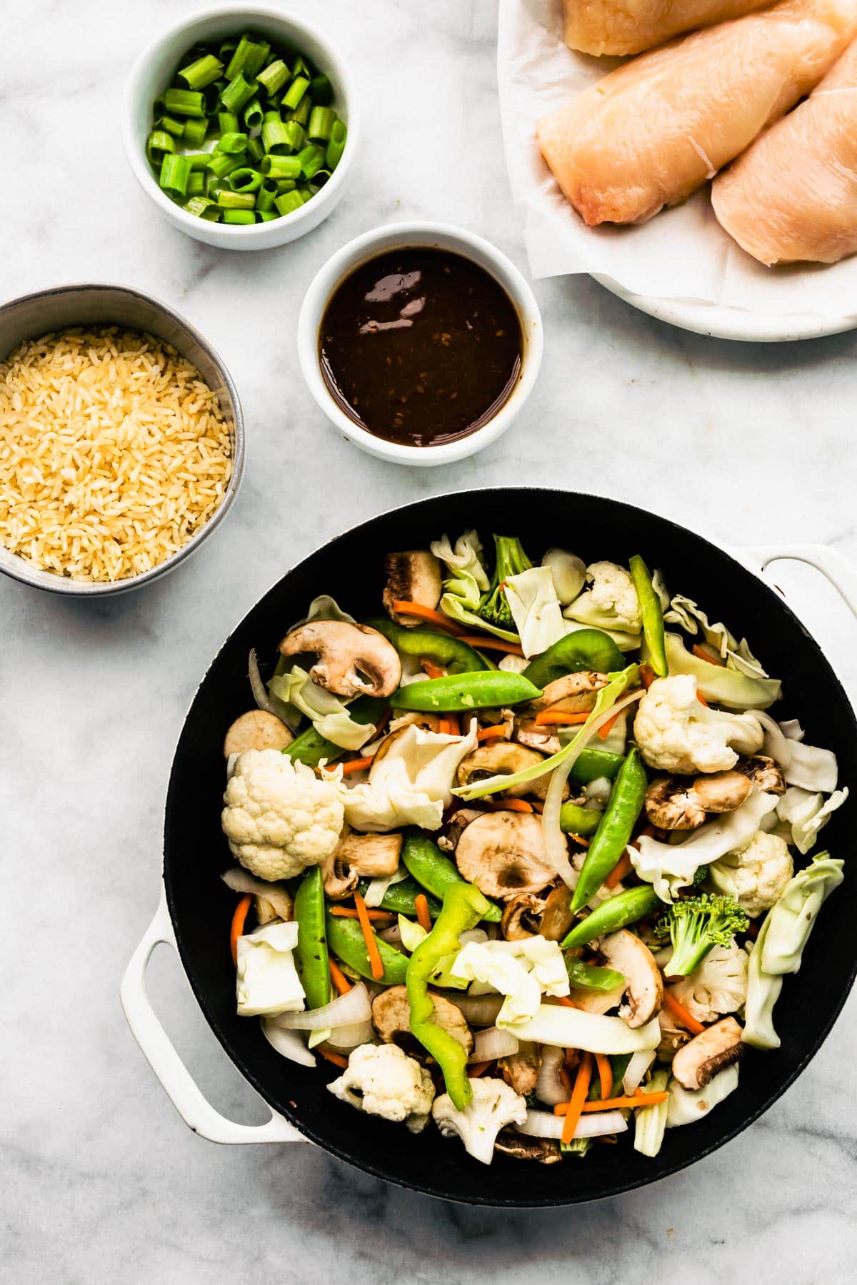 Asian vegetables, rice, chicken breasts and teriyaki sauce in bowls on a marble countertop.