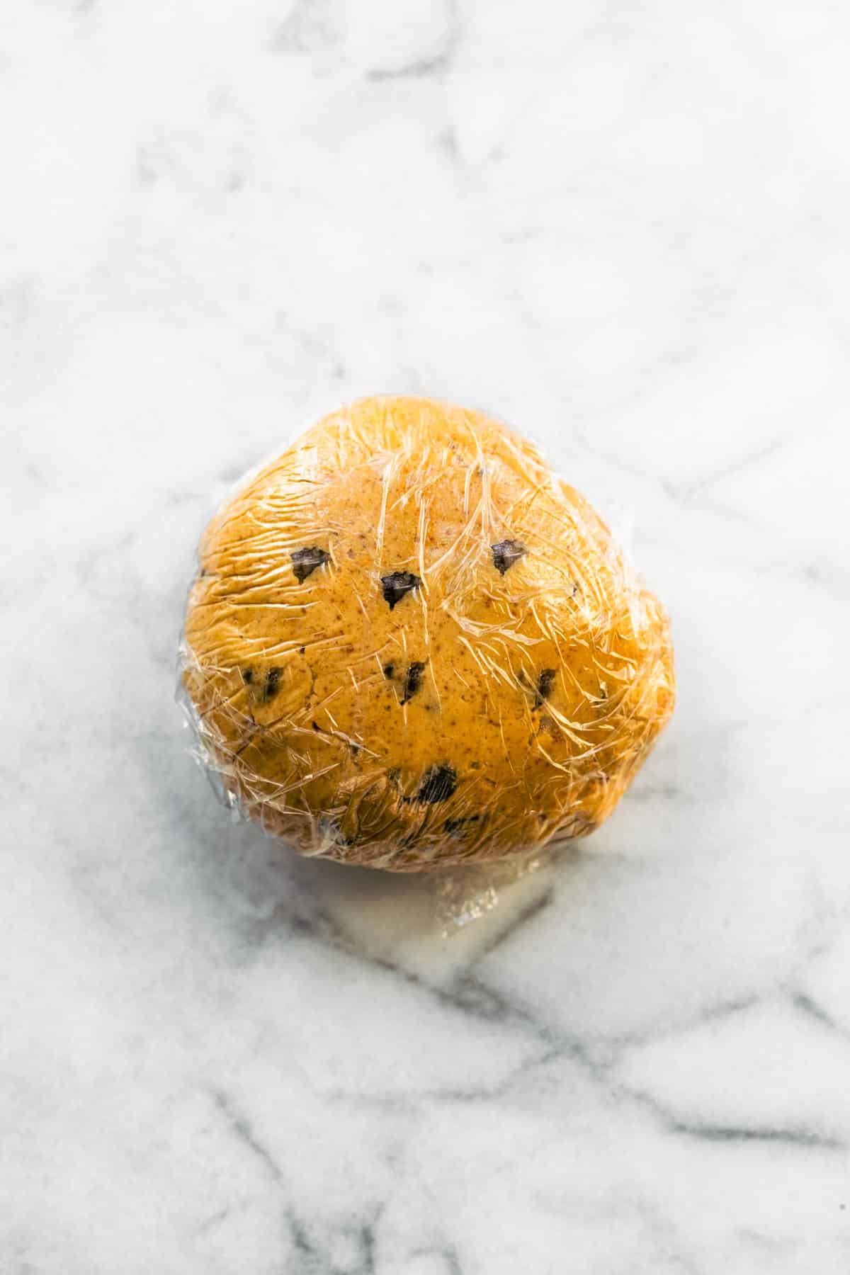 A ball of chocolate chip cookie dough wrapped in plastic wrap on a marble countertop.