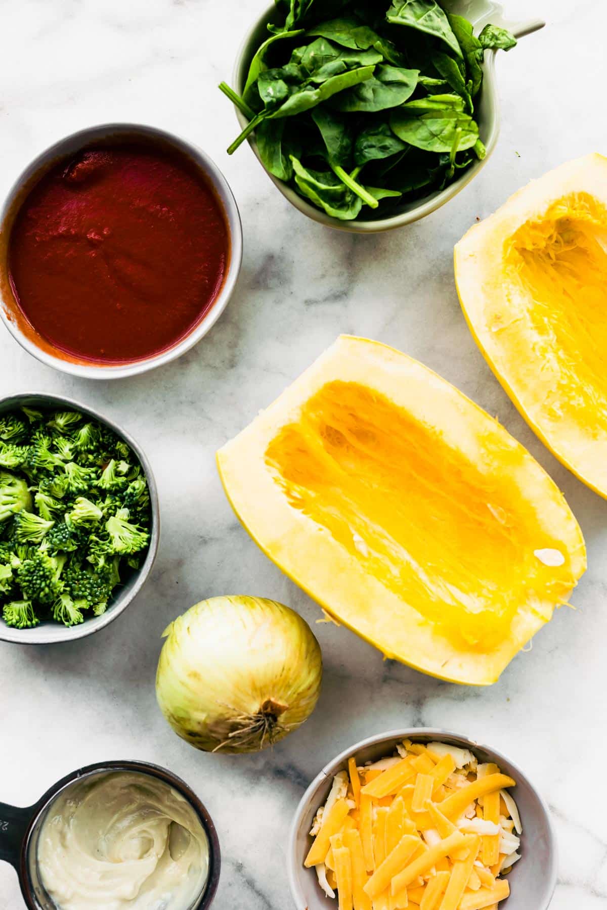 Halved spaghetti squash, marinara sauce, spinach and other ingredients on a white marble countertop.