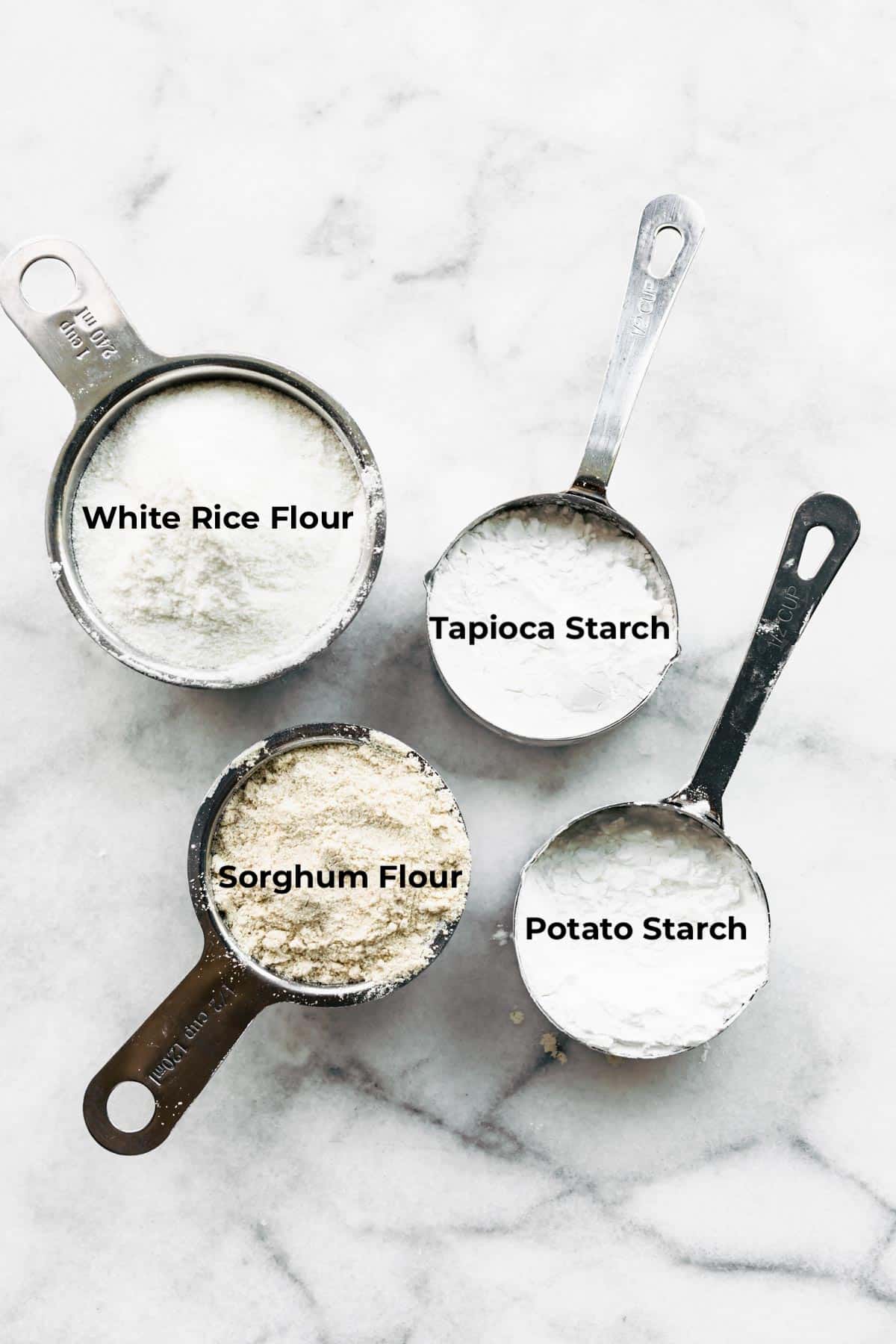 Four measuring cups on a marble countertop filled with different gluten free flours and labeled.