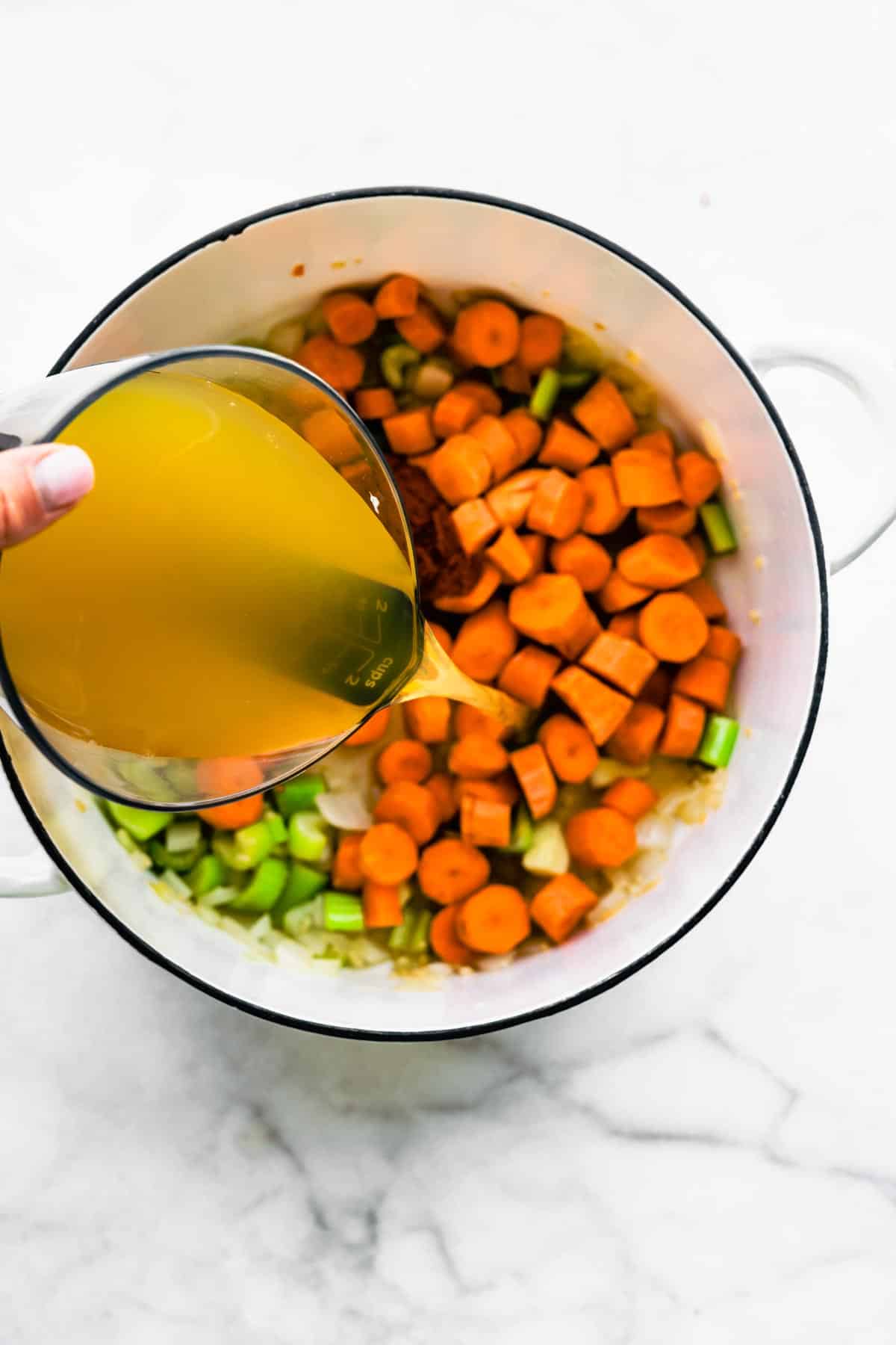 Vegetable broth being poured into a soup pot of diced carrots and celery.