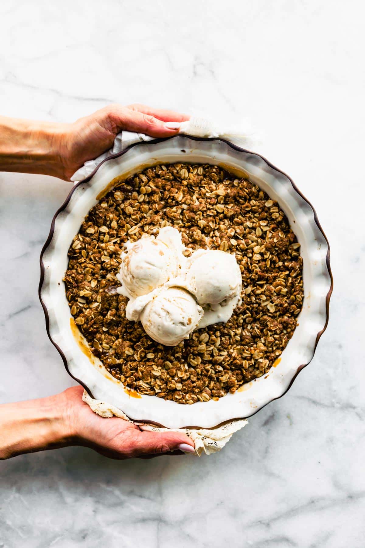 Overhead photo of two hands holding an apple crisp topped with vanilla ice cream.
