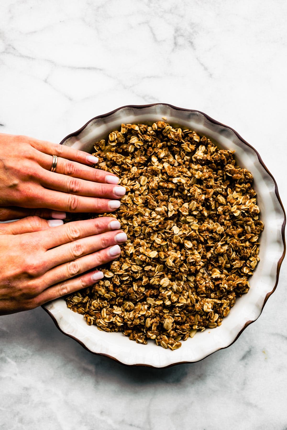 Two woman's hands pressing gluten free oatmeal crumble into a pie pan of apple crisp.