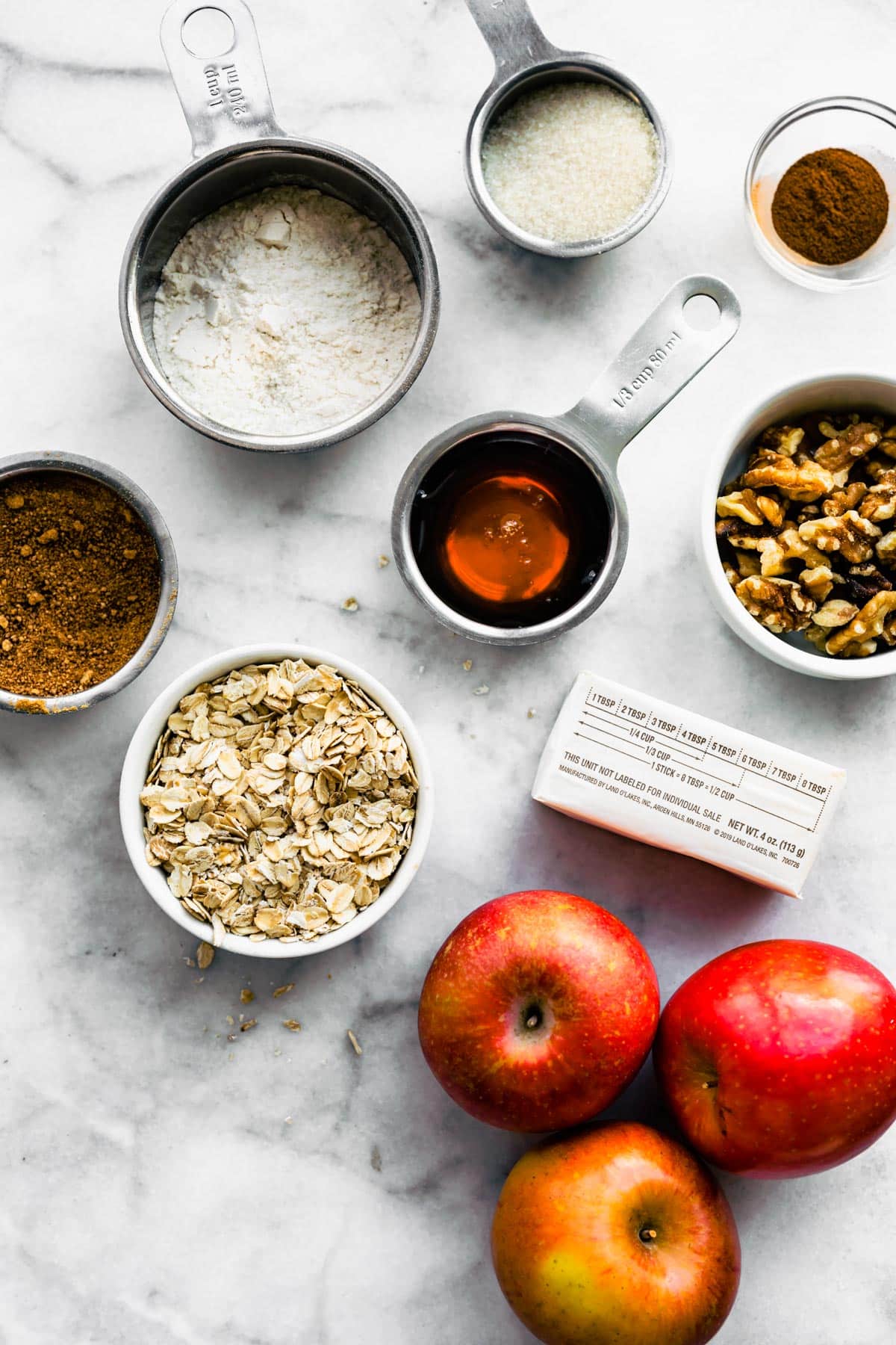 Red apples, oatmeal, maple syrup, butter and other ingredients on a white marble countertop.