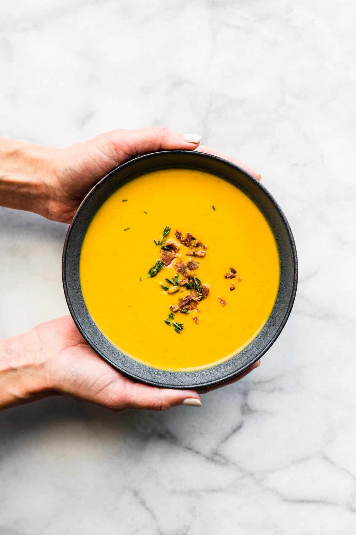 Two woman's hands holding a bowl of butternut squash soup topped with fresh thyme.