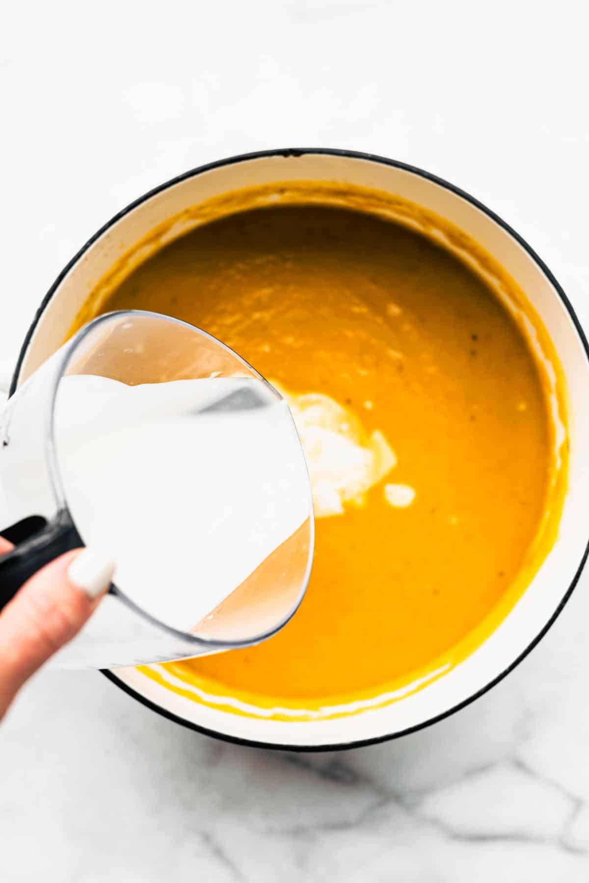 Creamy coconut milk being poured into a pot of vegan butternut squash soup.