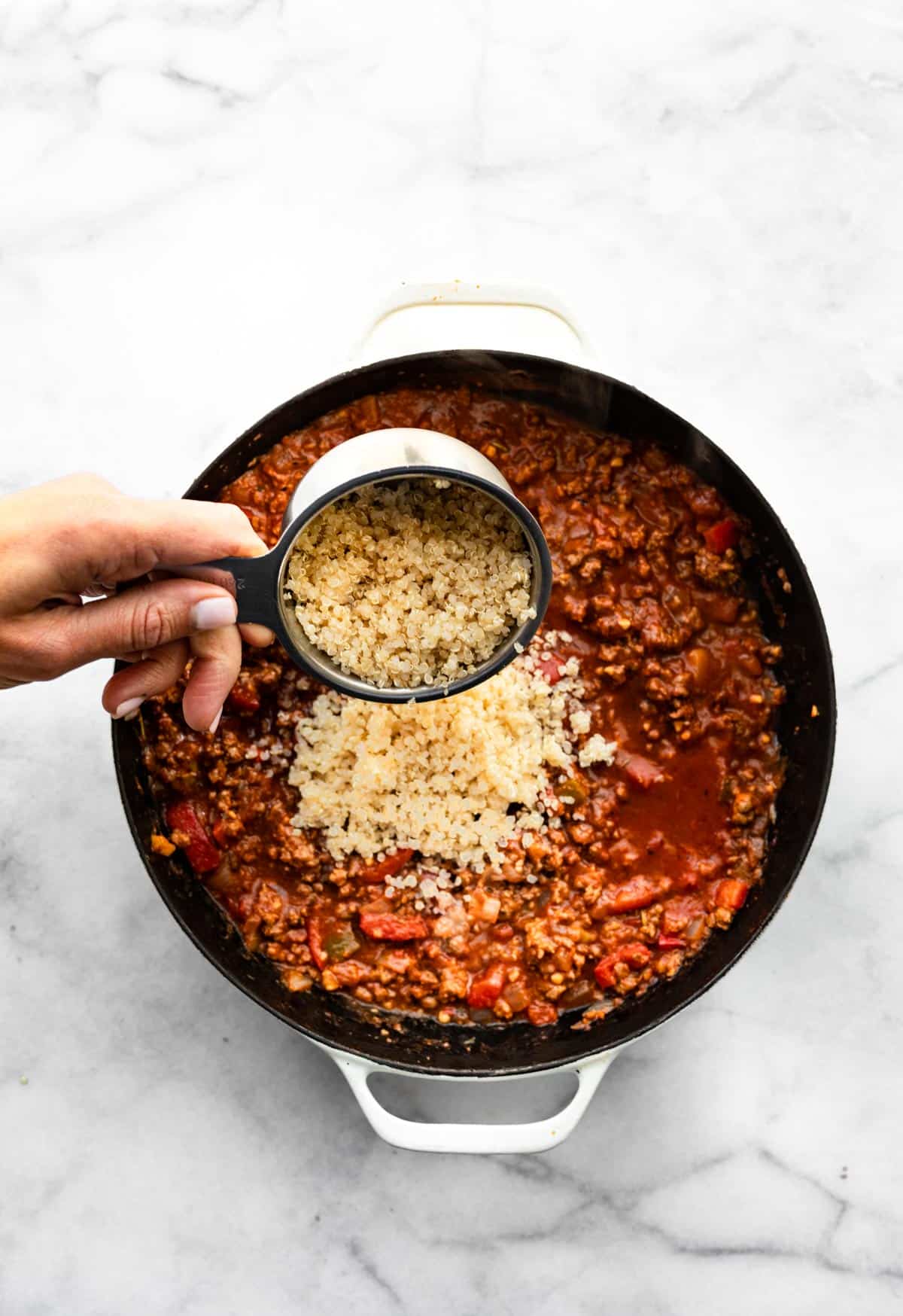 Cooked quinoa being poured into a pan of tomato based ground beef and peppers.