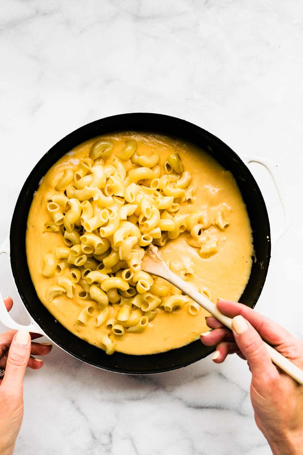 A woman's hand holding a wooden spoon stirring gluten free noodles into a cheese sauce.