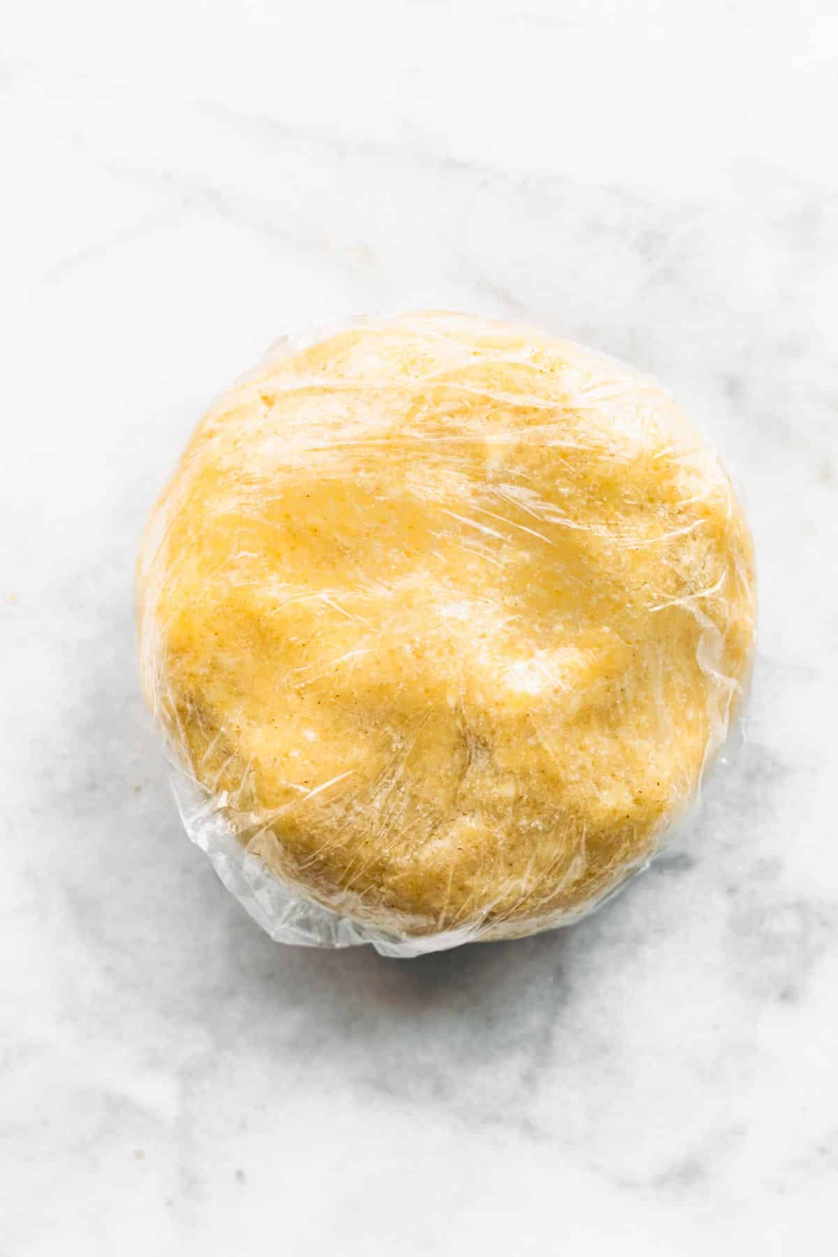 A gluten free pie dough puck wrapped in plastic wrap on a marble countertop.
