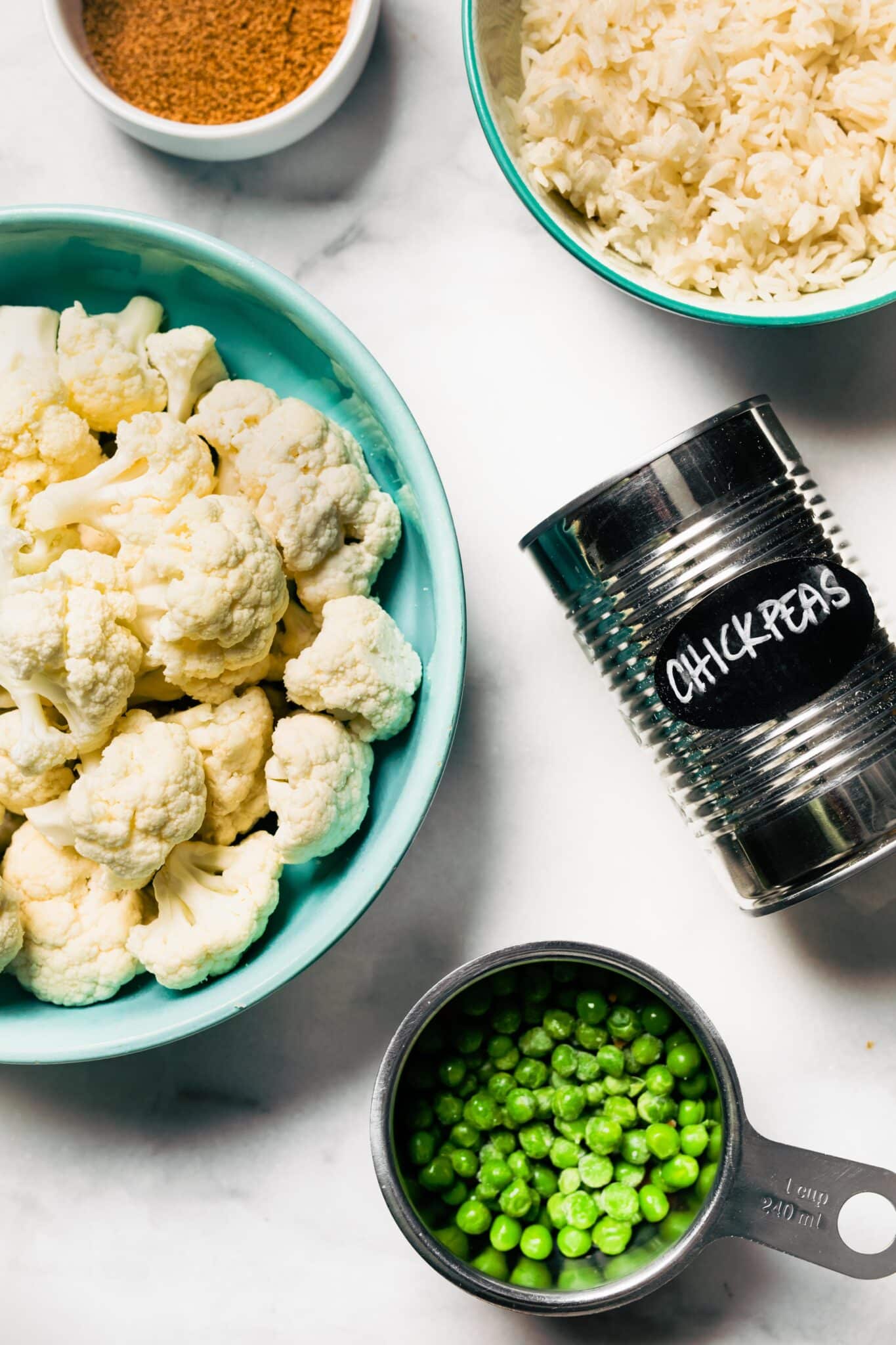 Bowls of cauliflower, rice, green peas and a can of chickpeas on a white countertop.