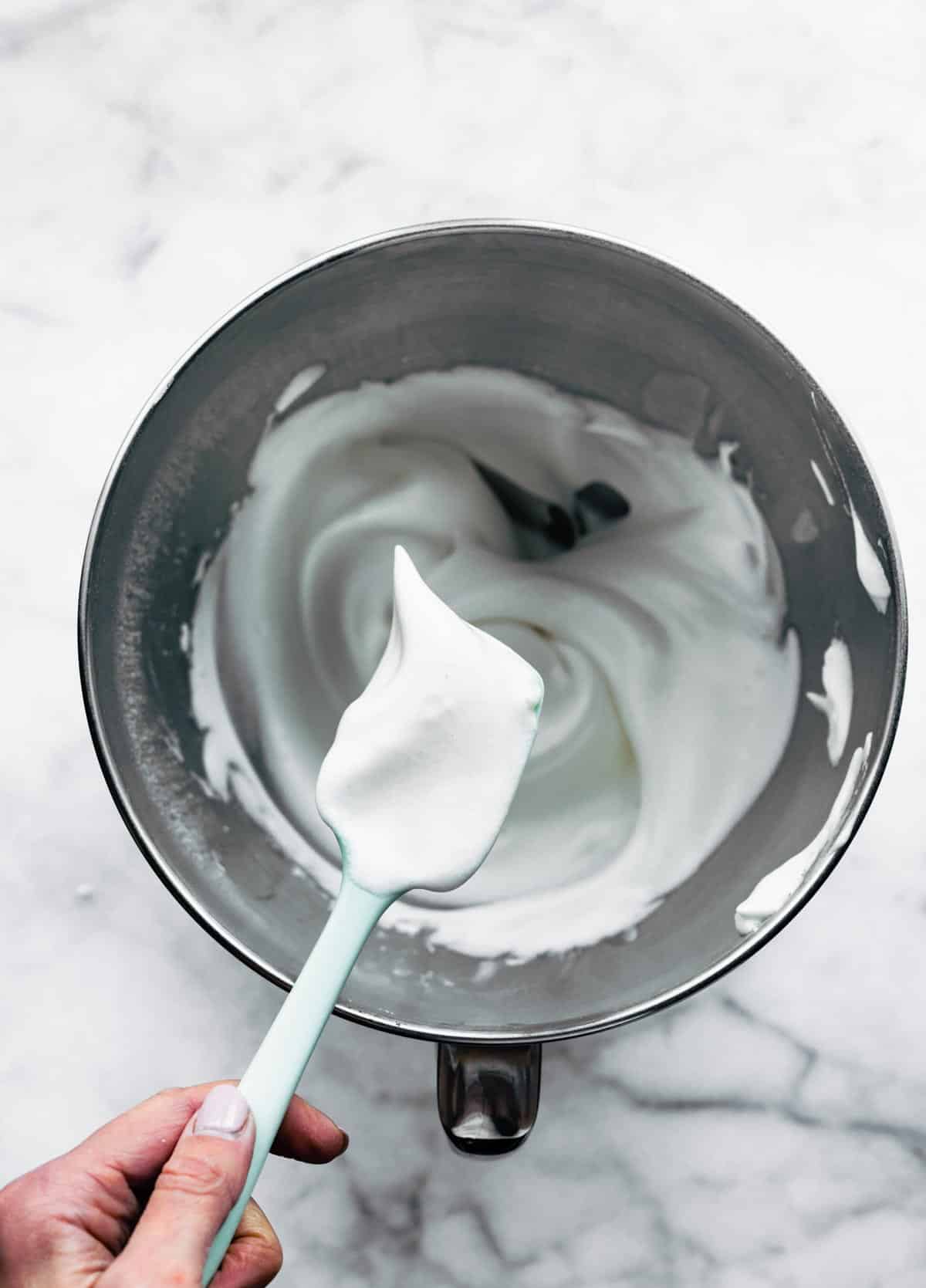 A woman's hand holding a spatula of whipped cream over a mixing bowl.