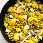A spoon digging into a cast iron pan of vegetarian curried cauliflower bake with chickpeas.