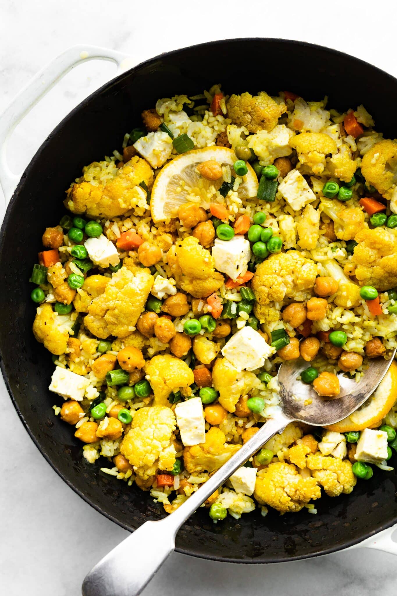 A spoon digging into a cast iron pan of vegetarian curried cauliflower bake with chickpeas.