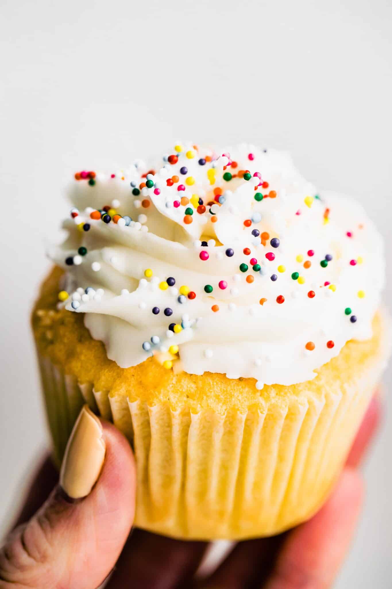 up close photo of woman's hand holding gluten free vanilla cupcake with sprinkles.