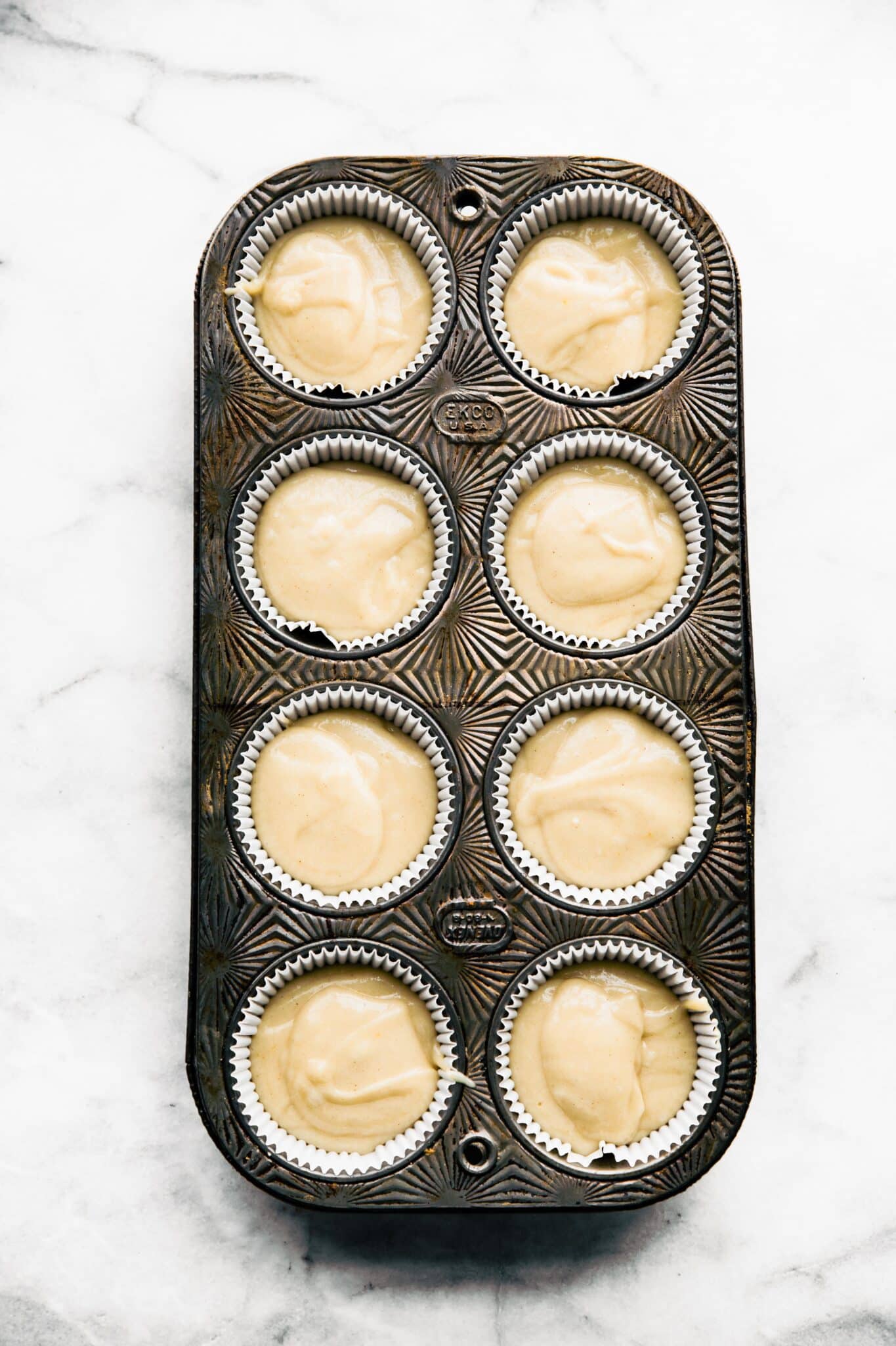Gluten free vanilla cupcake batter in 8 muffin liners in a vintage muffin tin.