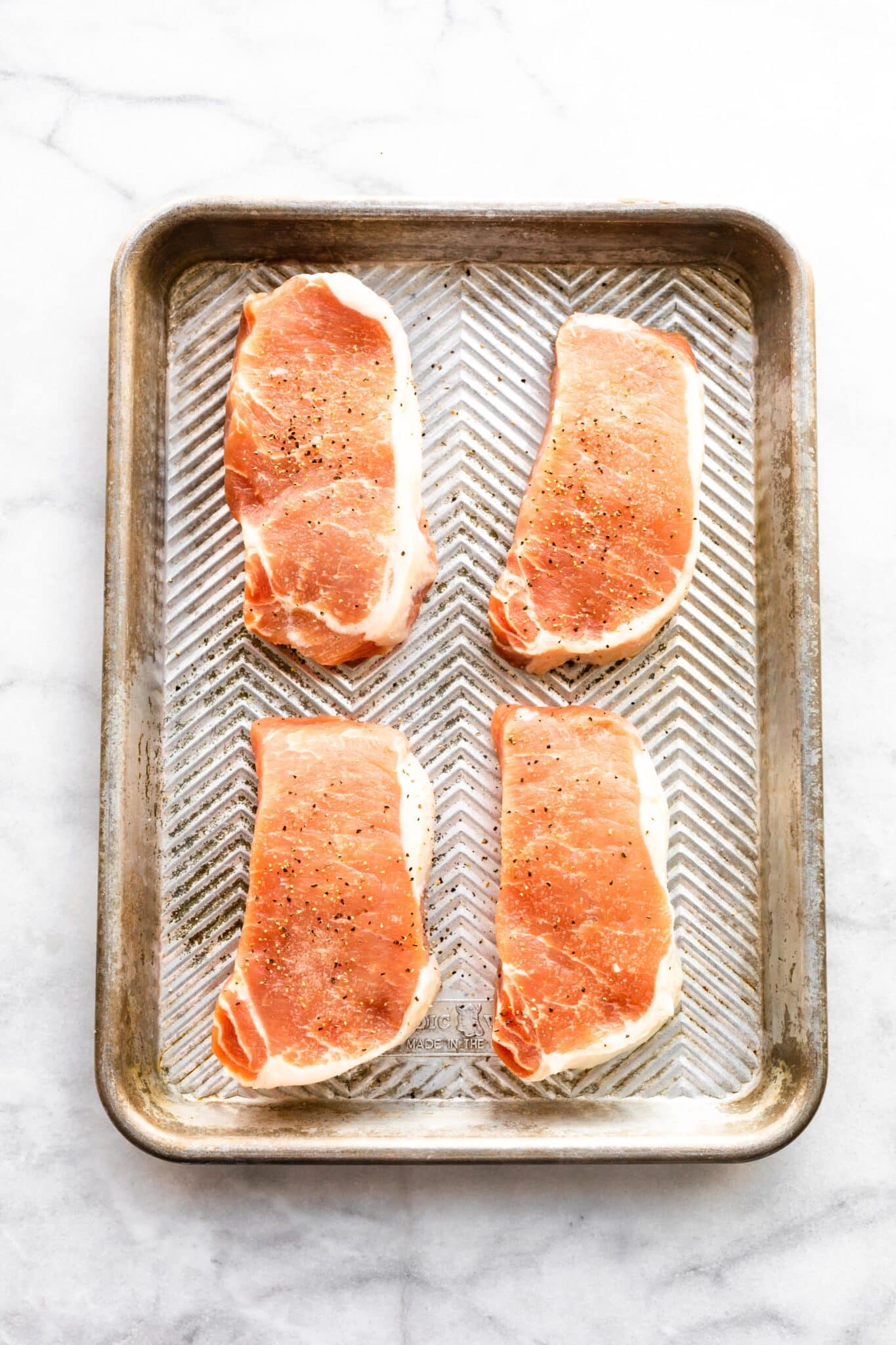 Four boneless pork chops seasoned with salt and pepper on a metal sheet pan.