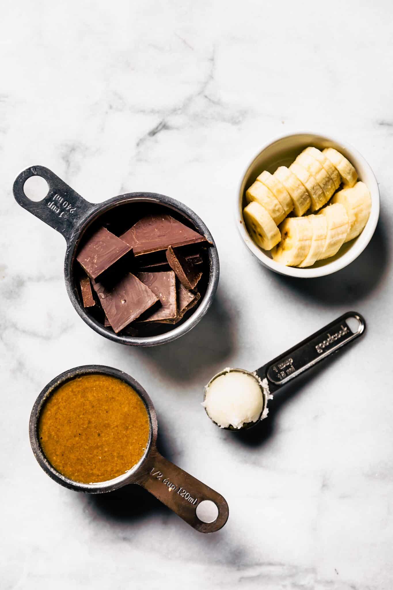 Dark chocolate pieces, banana slices, almond butter and coconut oil in bowls and measuring cups.
