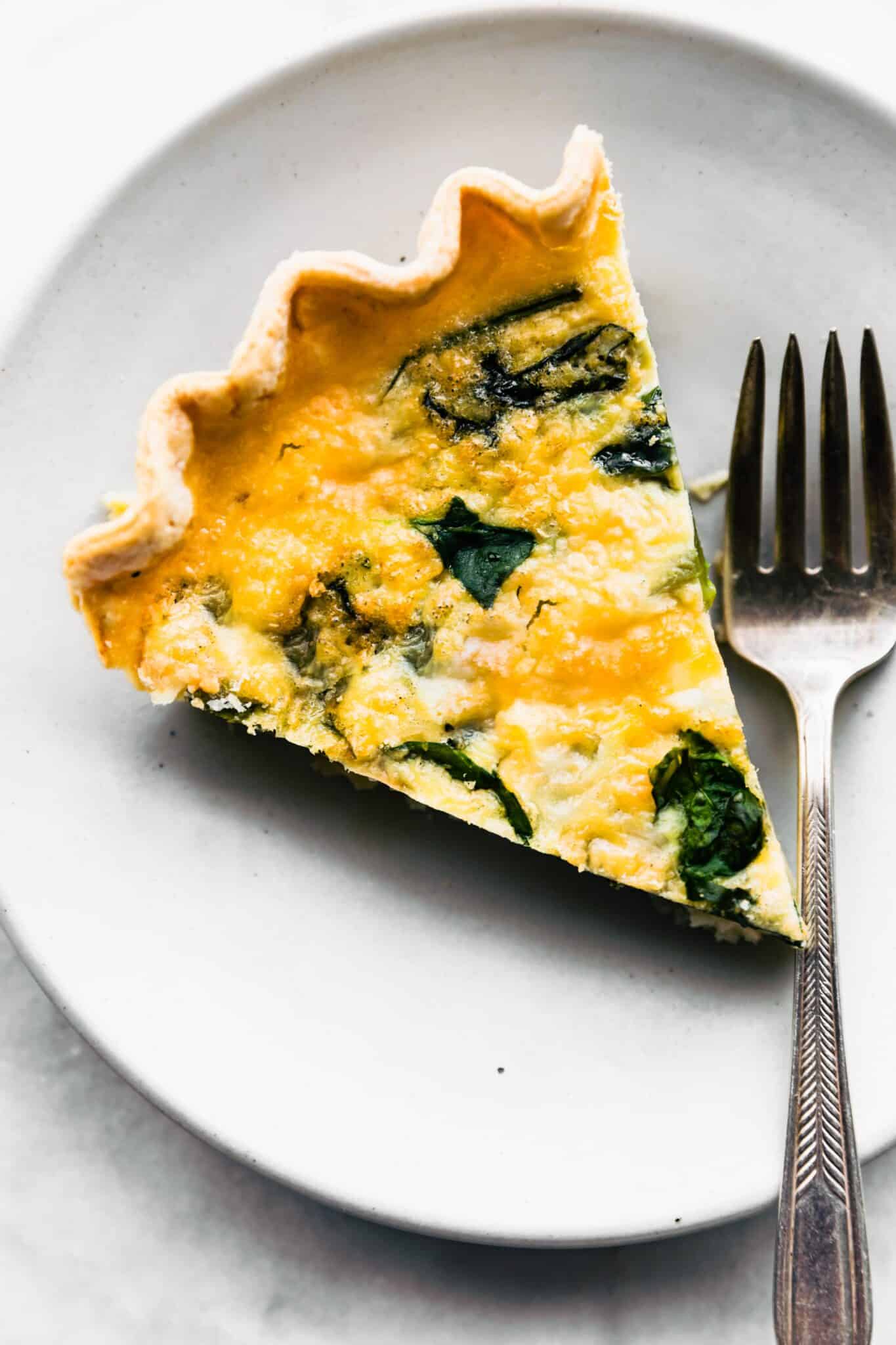 A slice of gluten free quiche on a plate with a fork beside it.