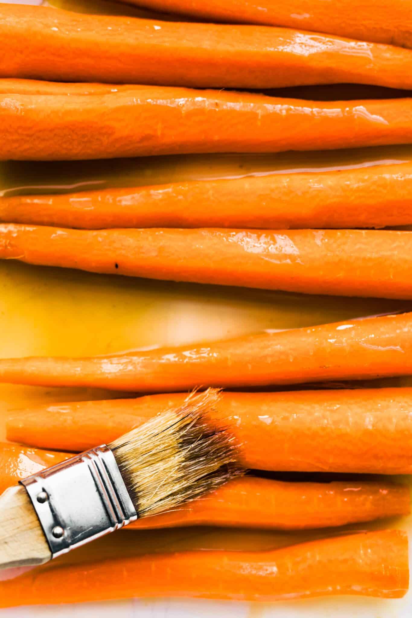 Up close photo of a brush glazing raw carrots with a maple bourbon glaze.