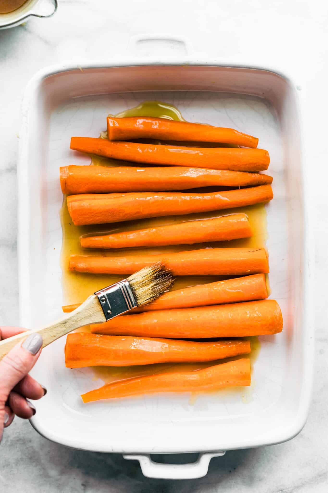 A woman's hand with a brush glazing raw carrots with a maple bourbon sauce.