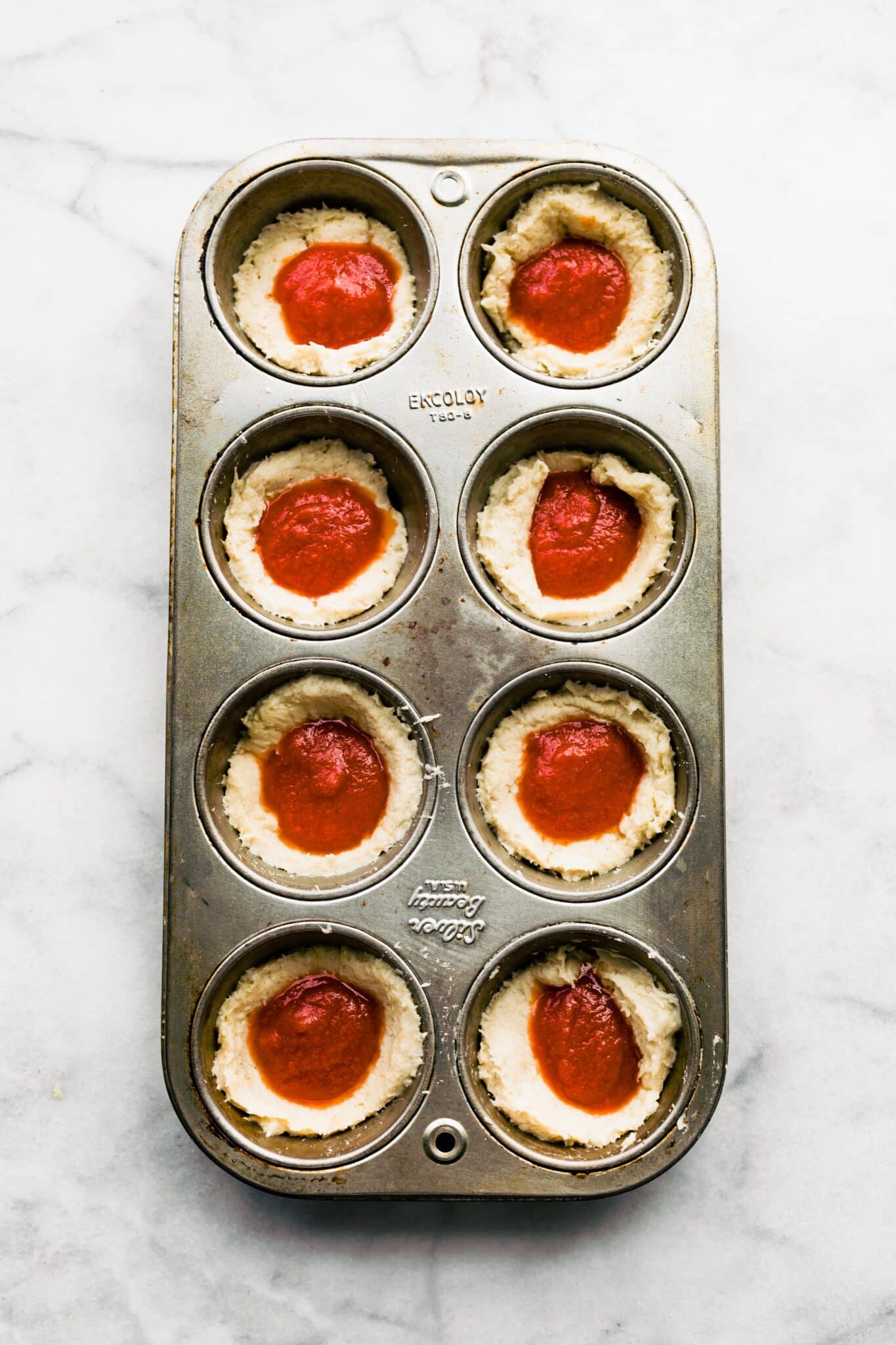 Overhead photo of an 8 count muffin tin with pizza dough and pizza sauce.