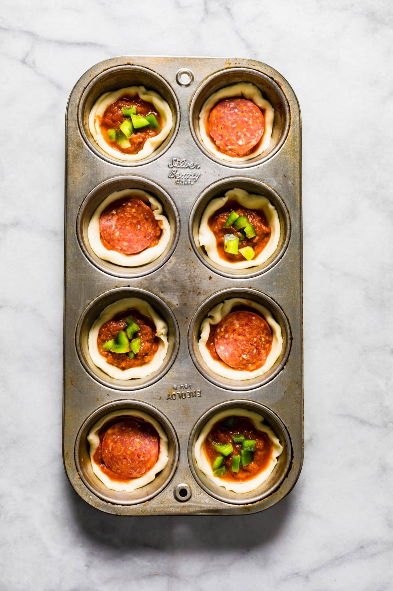 Overhead photo of an 8 count muffin tin with pizza dough, peppers, and pepperonis.