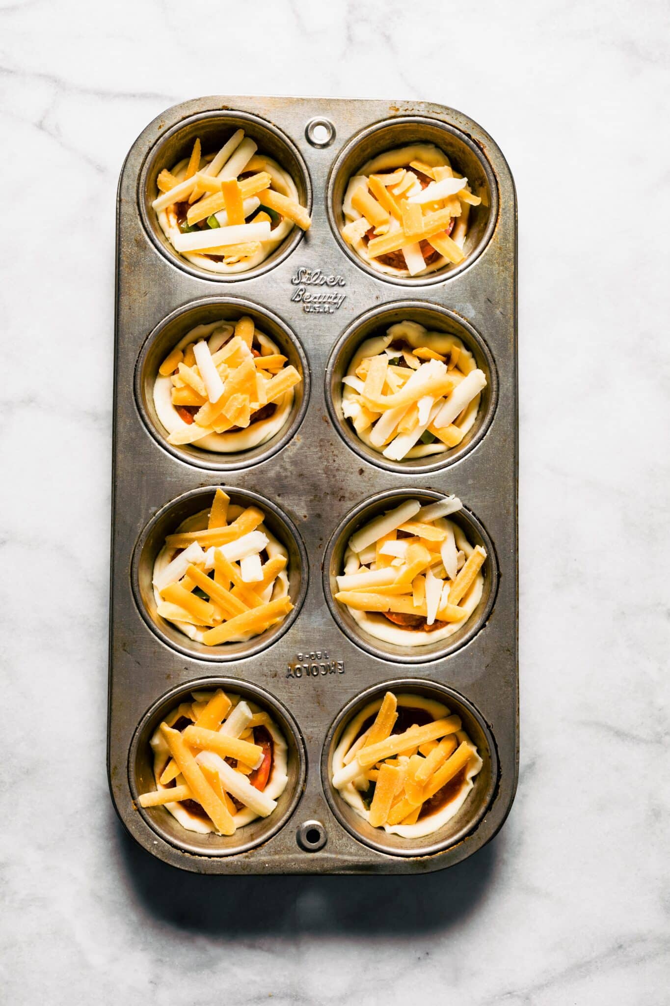 Overhead photo of an 8 count muffin tin with pizza bites topped with cheese.