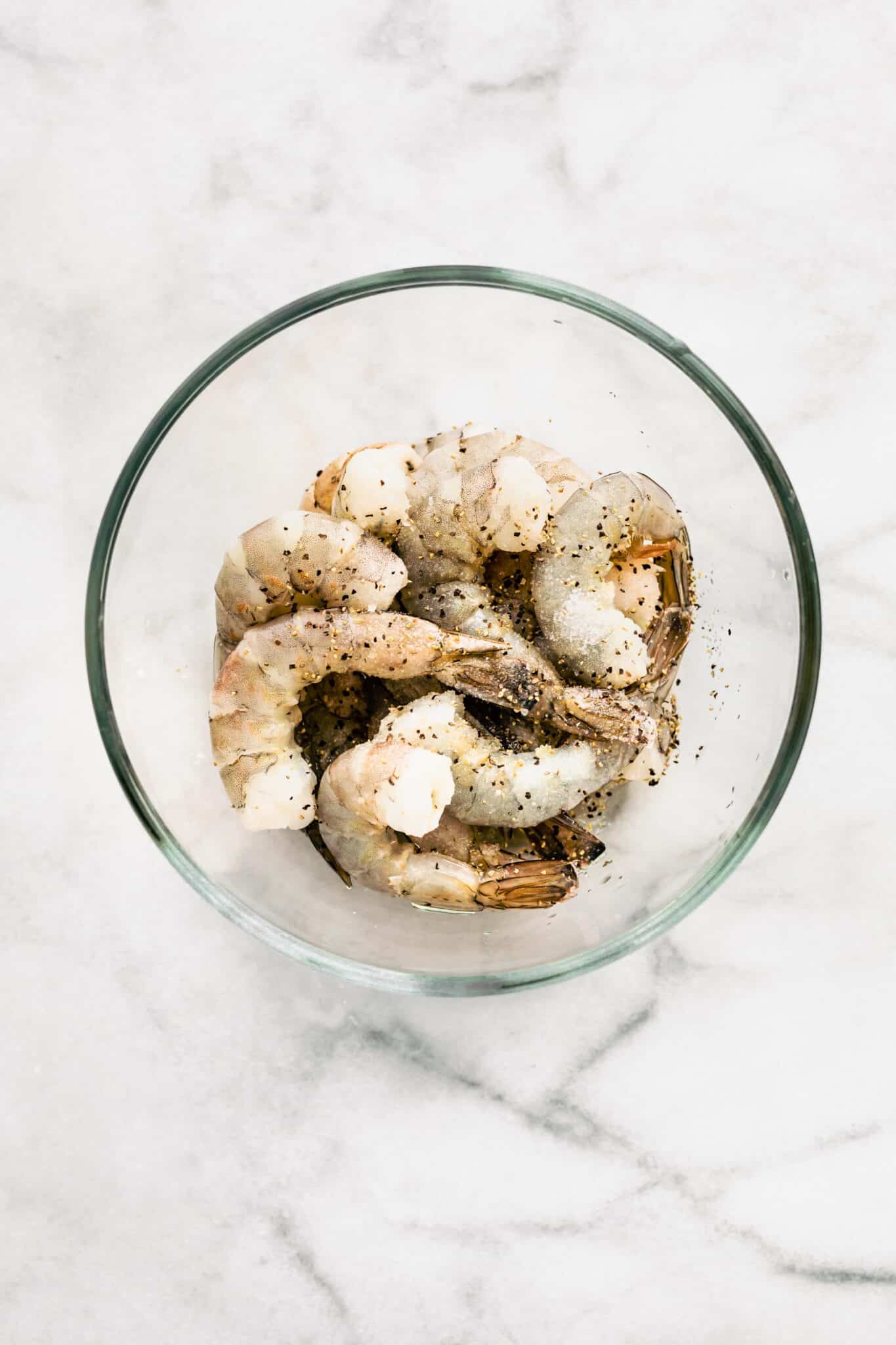 Overhead photo of raw shrimp sprinkled with salt and black pepper in a glass bowl.