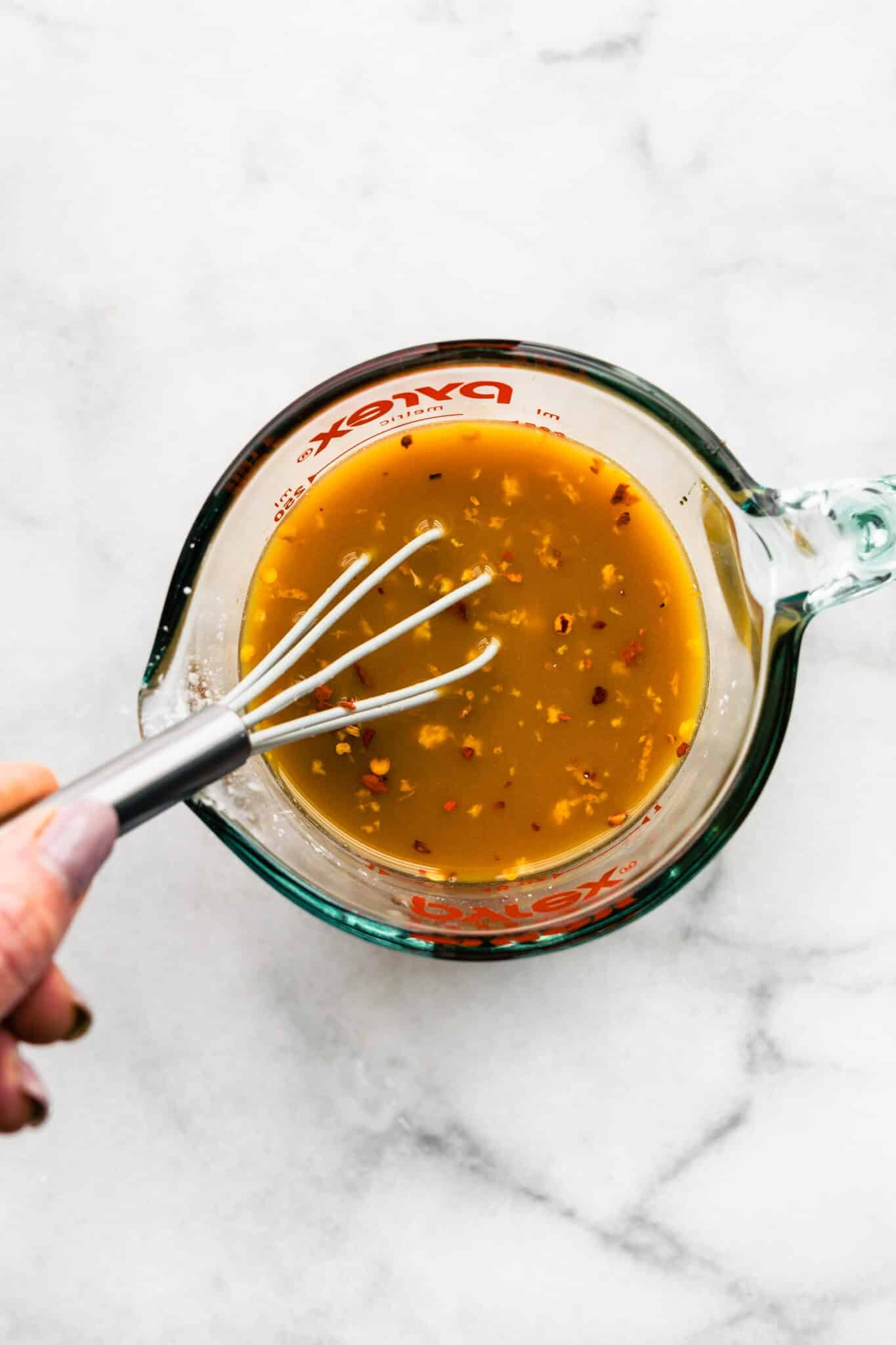 A whisk in a woman's hand whisking orange sauce in a glass measuring cup.