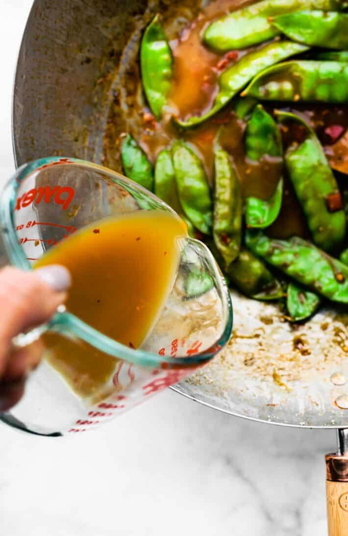 A woman's hand pouring orange sauce into a wok of sauteed green beans.